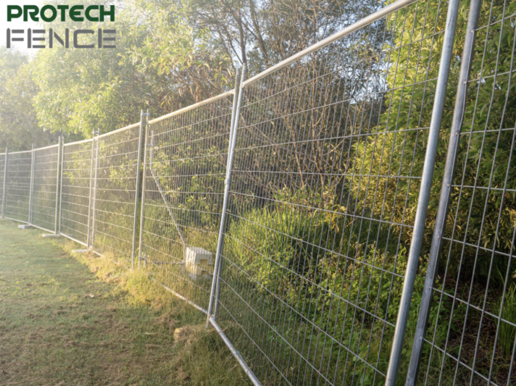A series of metal temporary fence panels with a grid pattern stand along a grassy pathway, bordering dense greenery in a sunlit outdoor area, providing a secure and temporary perimeter.