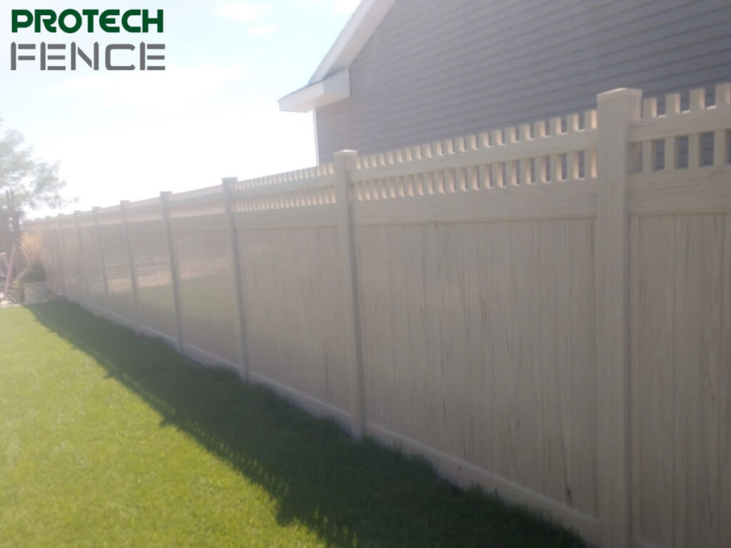 A 12 foot vinyl fence extends across the image. The fence is cream-colored with vertical panels and a decorative lattice top. The bright sun casts a sharp shadow of the fence onto the well-kept green lawn beside a residential building.