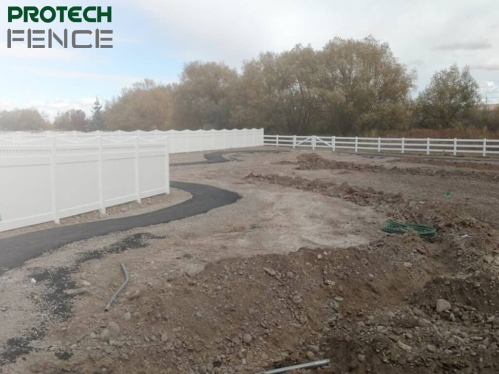An image showcasing a newly installed 7 foot vinyl fence by Protech Fence. The fence is pristine white with a scalloped top design, sectioning off a landscaped area with a curved pathway and contrasting against a backdrop of trees and an overcast sky. The foreground shows an under-construction area with scattered dirt and construction materials.