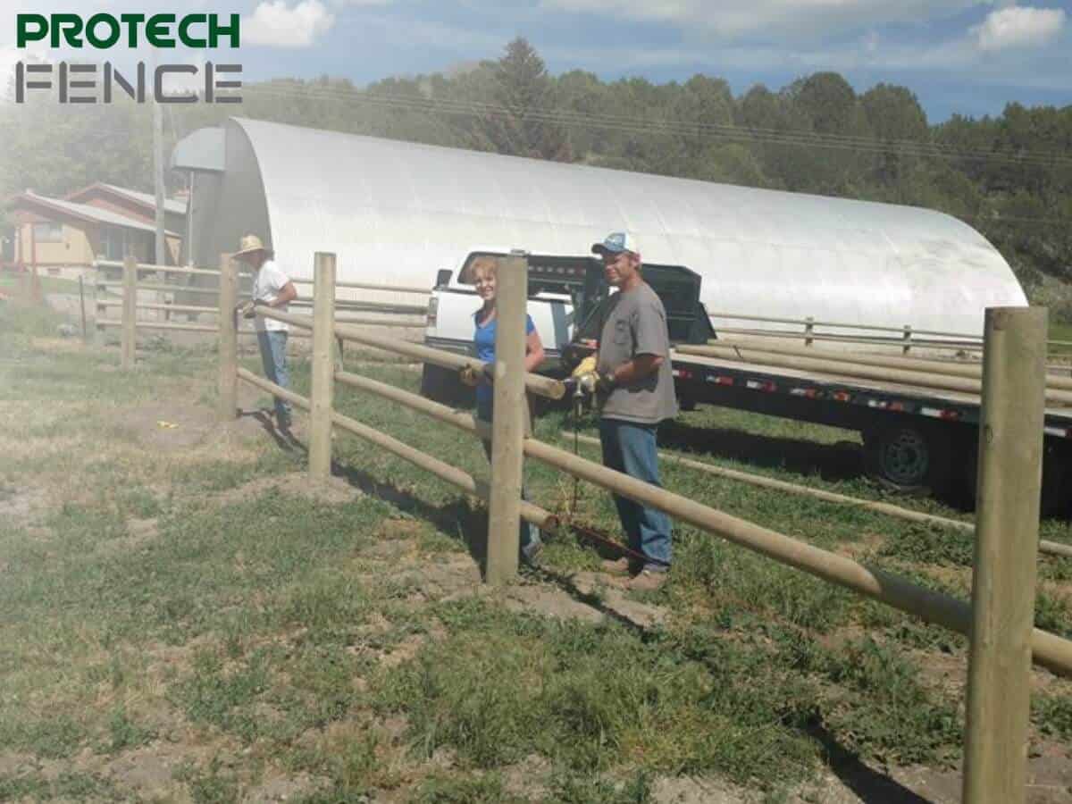 Two Protech Fence employees working on a wooden fence repair in Pocatello, with one holding a drill and the other assisting, in a field with lush green trees in the background.