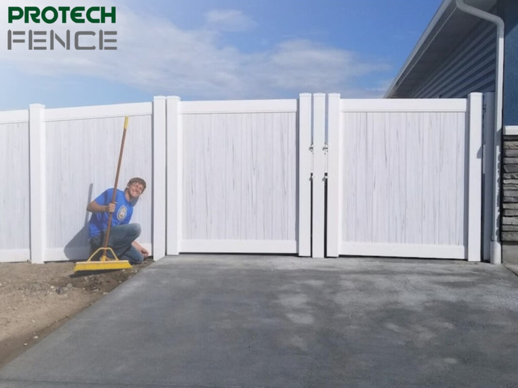 A smiling worker from fence contractors Pocatello using a yellow level to ensure the alignment of a newly installed white vinyl fence next to a modern house with a stone facade and a grey concrete driveway.