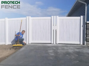 A smiling worker from fence contractors Pocatello using a yellow level to ensure the alignment of a newly installed white vinyl fence next to a modern house with a stone facade and a grey concrete driveway.