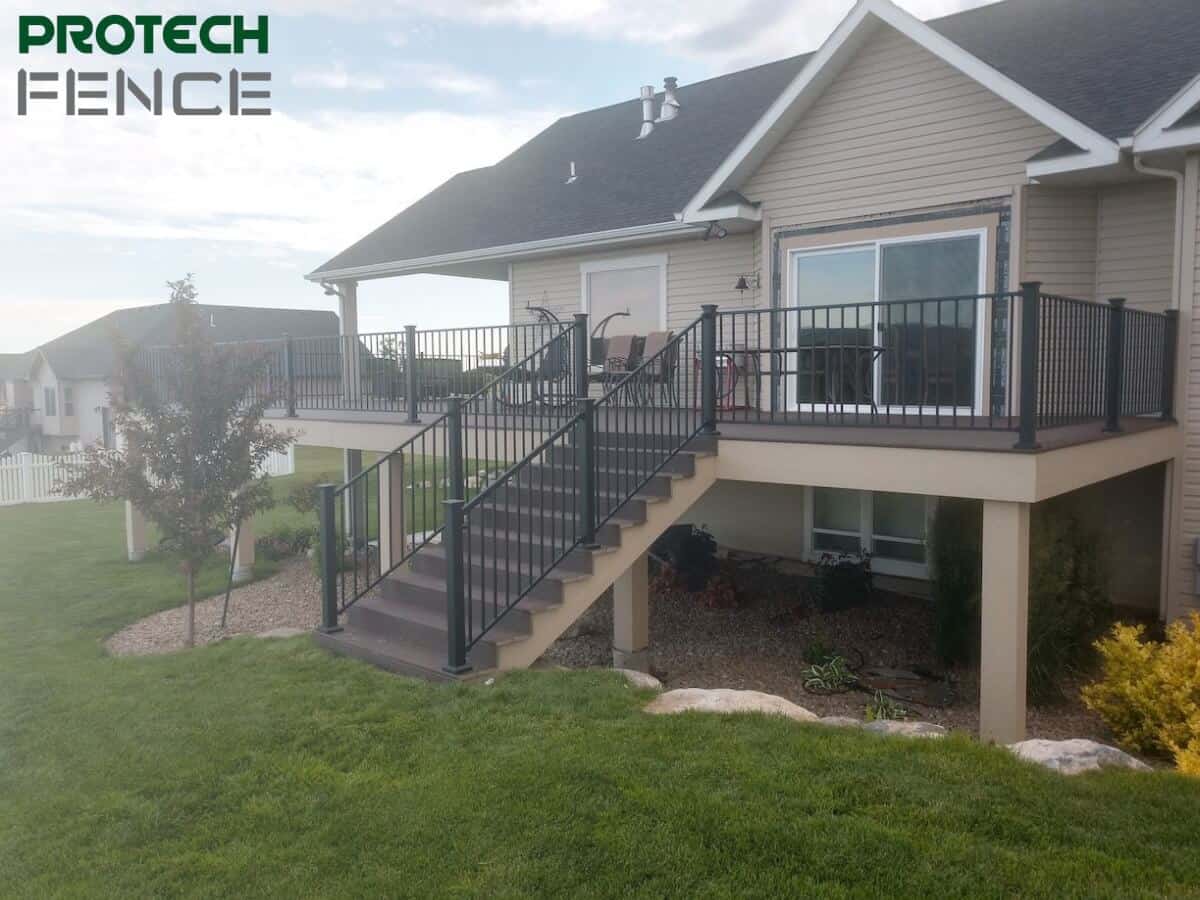 Elevated beige deck with dark railing installed by deck contractors Pocatello, featuring steps and landscaped garden, enhancing a suburban home's backyard with the Protech Fence logo. 