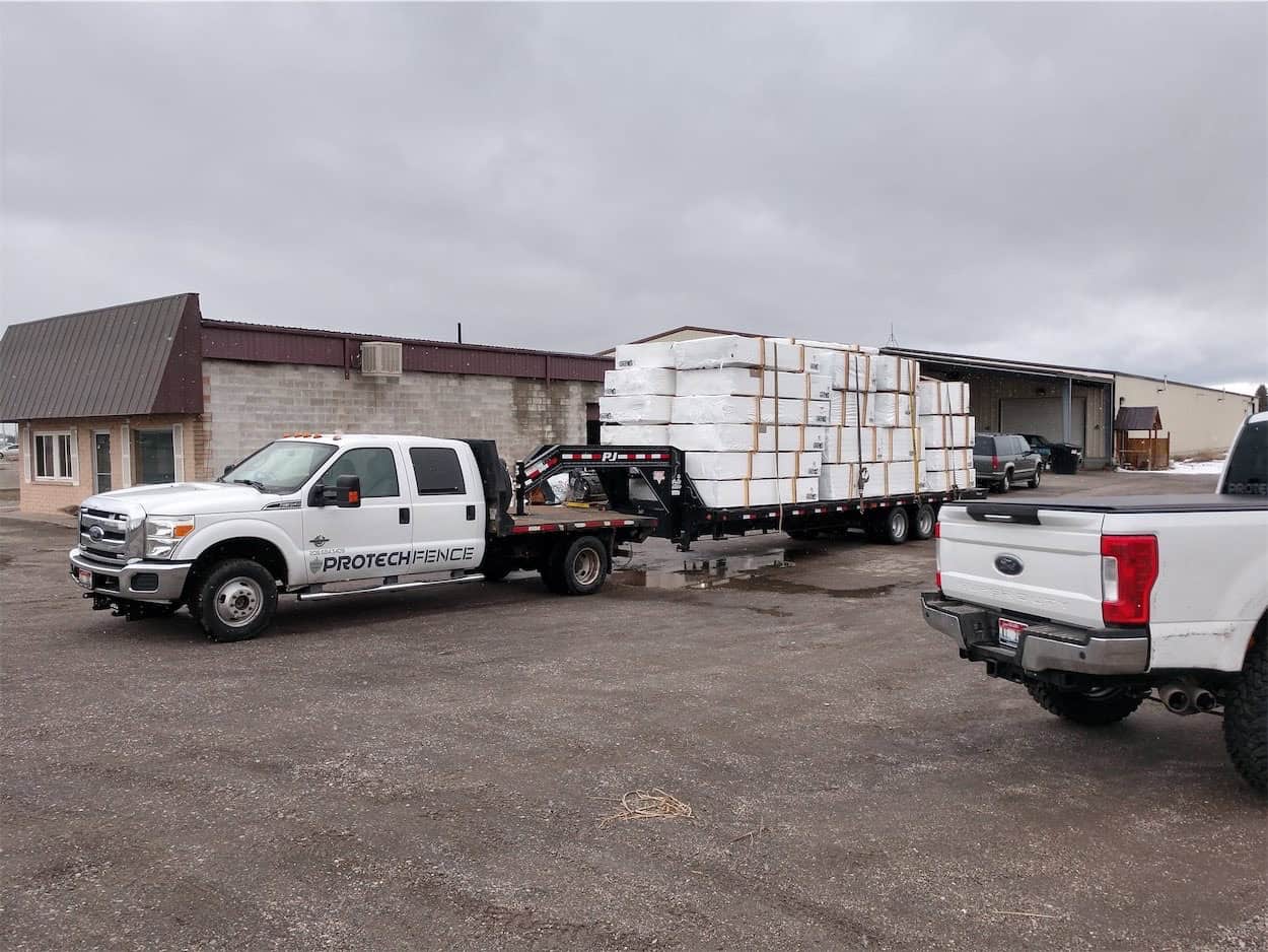 A Protech Fence branded white pickup truck is towing a trailer loaded with secured packages of fence supplies in Pocatello, stationed in an industrial area with buildings in the background, showcasing the company's readiness for delivery. 