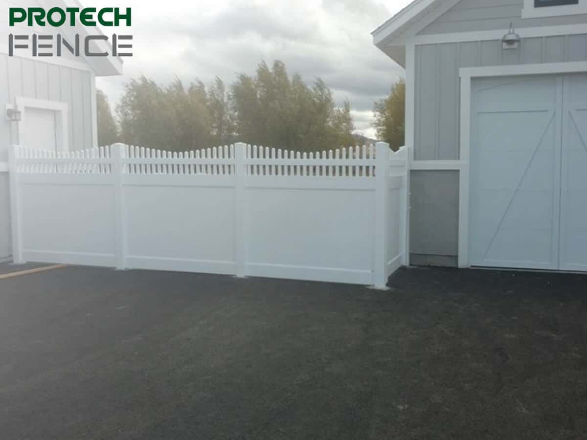 A 7 foot vinyl fence with a pointed picket design installed adjacent to a white garage, enhancing the property's privacy and curb appeal. The clean lines of the fence complement the garage's architecture, set against a gravel ground and soft natural backdrop. 