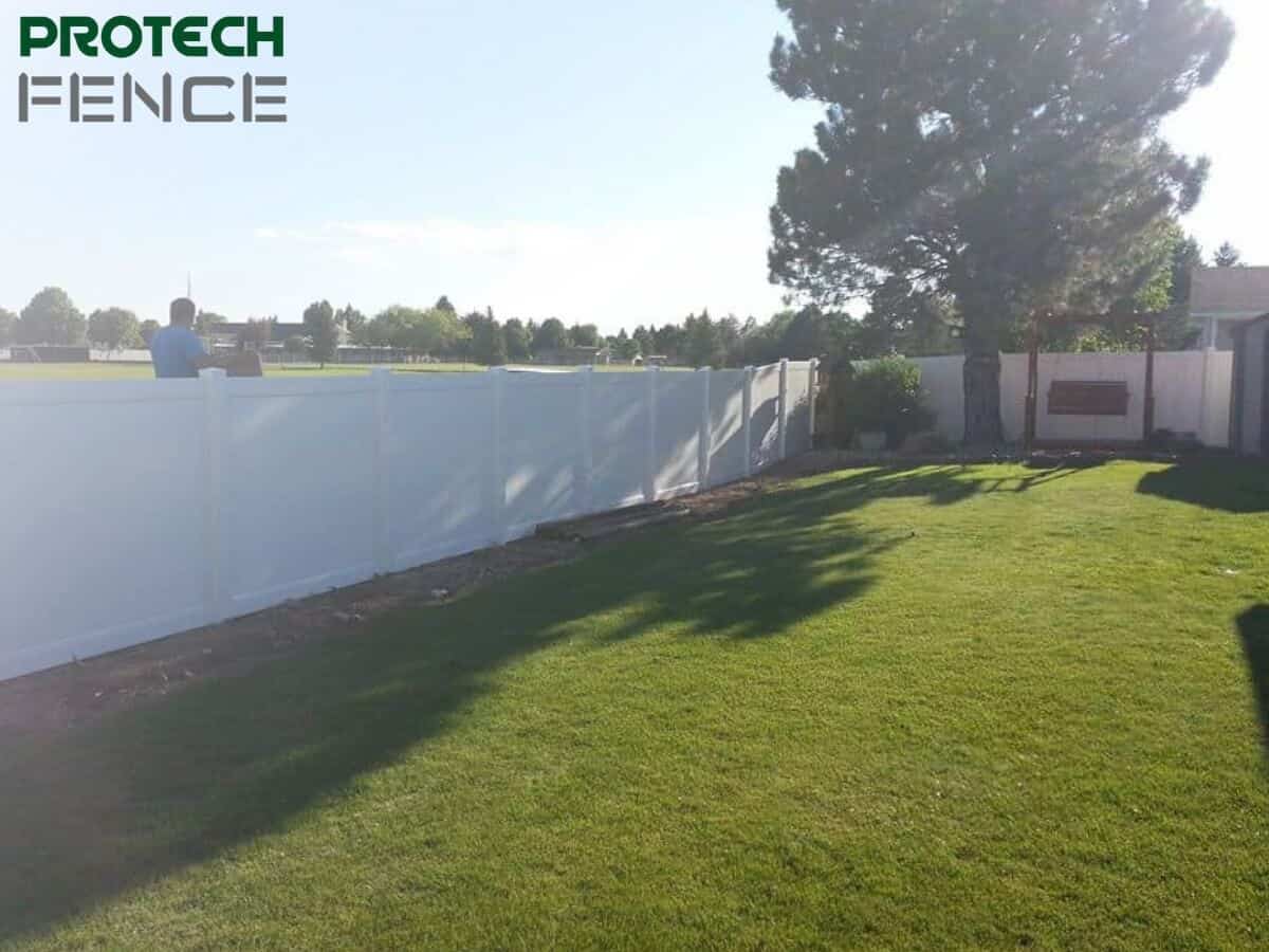 A person is seen from behind, working on a tall, pristine white fence in a lush backyard, indicative of the services provided by fencing companies that offer financing in Pocatello. The clear sky and the well-maintained grass suggest a serene suburban environment, perfect for privacy and home improvement projects. 