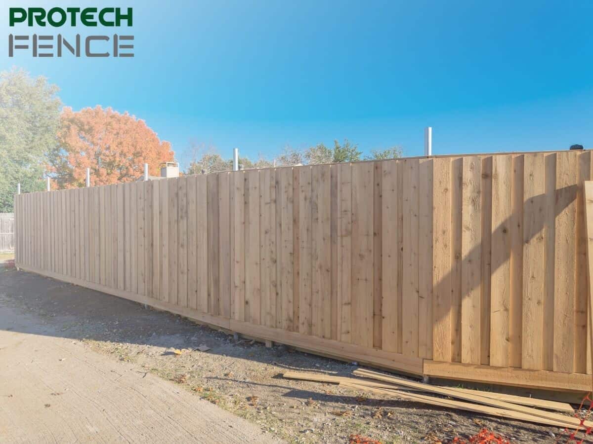 A new, seamless wooden privacy fence built by one of the fence companies in Pocatello, showcasing quality craftsmanship with the Protech Fence logo in the upper left corner, set against a clear blue sky and autumn foliage. 