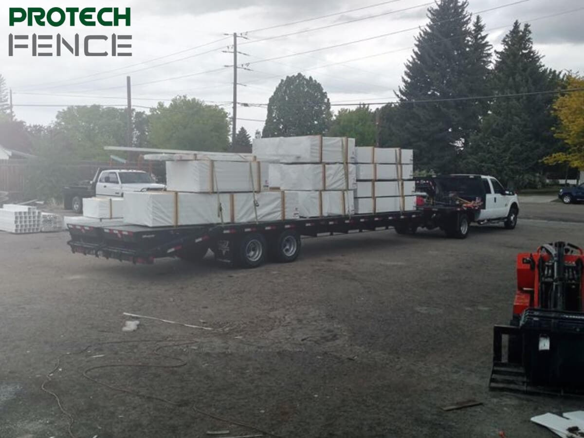 A flatbed trailer hitched to a pickup truck is loaded with neatly stacked and packaged fence supplies in Pocatello, ready for transport to a construction site, with a forklift in the foreground indicating ongoing loading operations. 