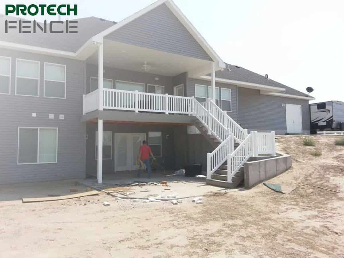 A worker from fence contractors Pocatello is busy with construction tools near a two-story house with a new white deck and stairs, indicating ongoing outdoor work on a bright sunny day.