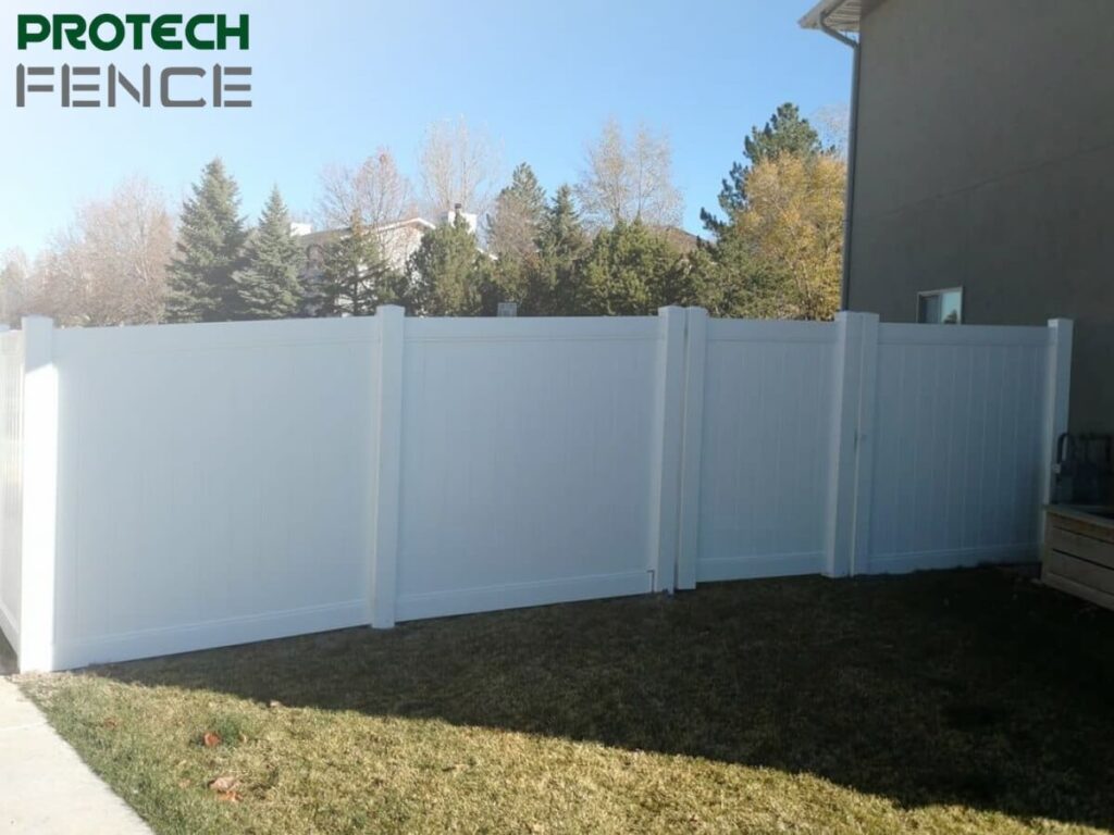A sturdy 5 ft vinyl fence gate installed in a suburban backyard with a solid white finish, flanked by matching vinyl fencing panels, on a sunny day with a clear blue sky and residential homes in the background.