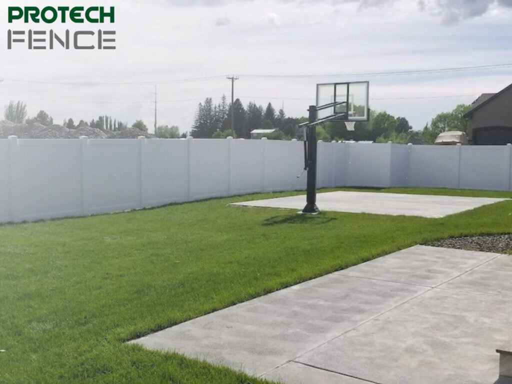 A serene backyard enclosed by a 10 ft vinyl fence gate. The landscape features a well-maintained lawn, a concrete patio area, and a basketball hoop, offering a private recreational space. The tall vinyl fencing provides a secure and secluded environment for outdoor activities.