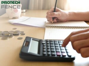 An individual is calculating finances at a wooden desk with a clear focus on budgeting for services. There is a piggy bank signifying savings, a calculator being used to crunch numbers, and a notepad where financial planning is being recorded. This scene represents the thoughtful planning of expenses and could be associated with the careful consideration of budgeting for home improvements, such as working with fencing companies that offer financing in Pocatello.