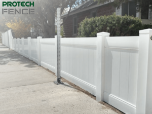A white vinyl fence lines the sidewalk beside a residential street, with a house partially visible behind the neat row of tall, solid panels and posts. The clear sky suggests a sunny day, ideal for showcasing the clean lines of the newly installed fencing. This visual would be relevant for discussions about vinyl fence installation cost per foot.