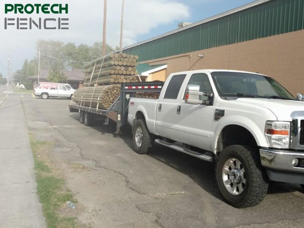 A white Protech Fence company truck parked outside a green industrial building, loaded with a stack of new, round fence posts for fence repair Pocatello services.