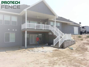 Deck contractors Pocatello working on a new deck installation at a residential home with a two-story gray siding exterior, featuring white railing and stairway, with the Protech Fence logo at the top