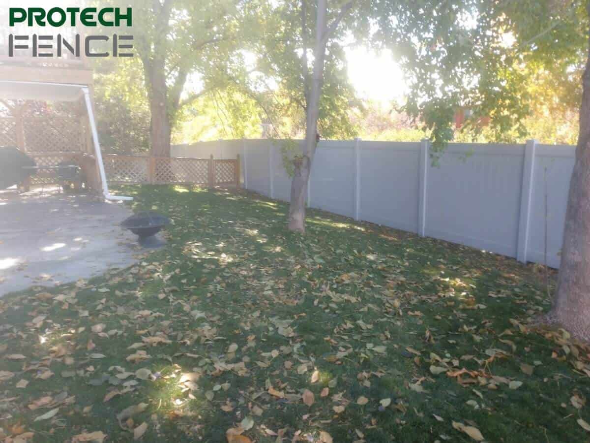 A tranquil backyard scene with a 5 ft vinyl privacy fence with a small open gate, displaying the Protech Fence logo, surrounded by trees with fallen leaves on the lawn, suggesting an autumn setting. 