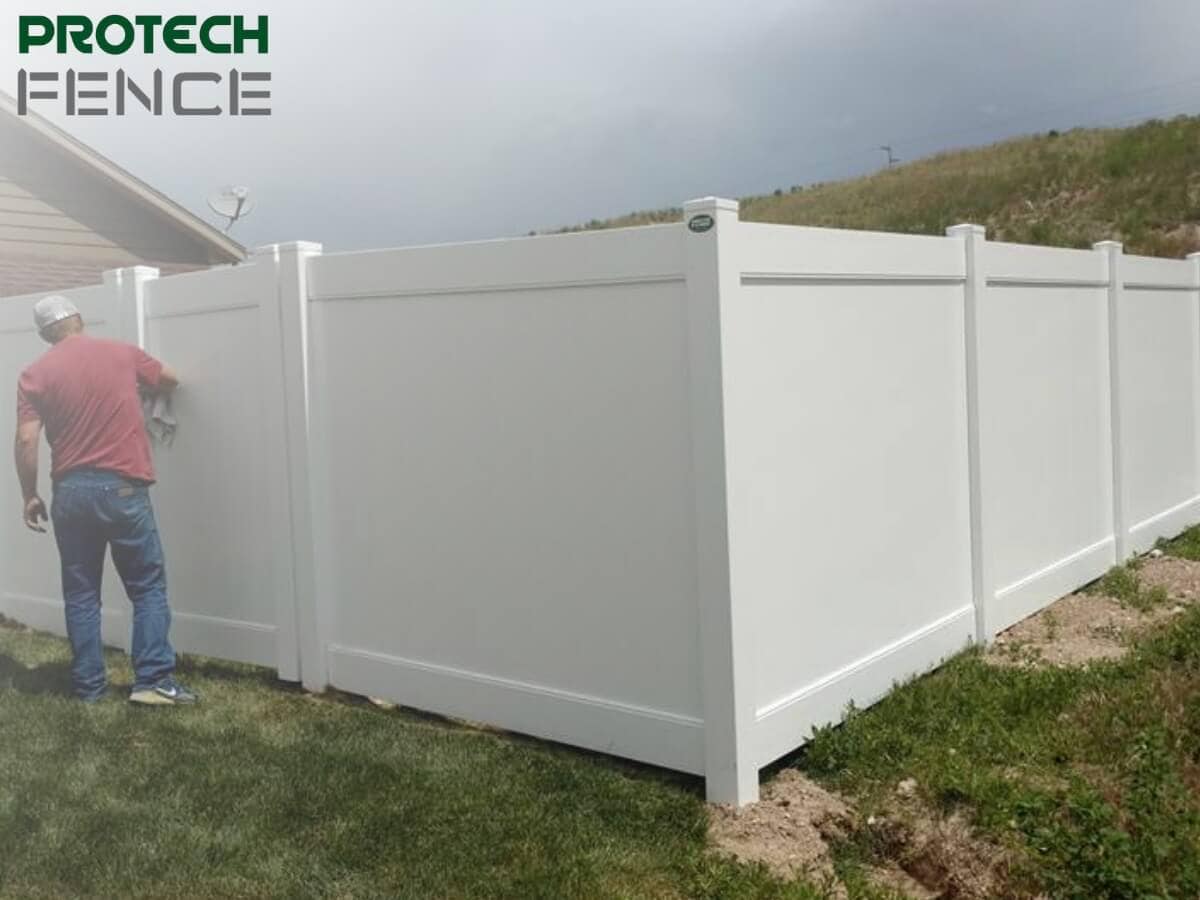 A fence contractor from Pocatello is seen applying finishing touches to a long, white vinyl privacy fence beside a residential home, under an overcast sky. 